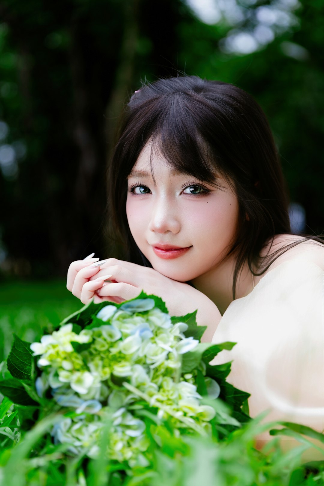 Smiling young woman lying on green grass with a bouquet of fresh hydrangea flowers, wearing a soft cream dress in natural light.